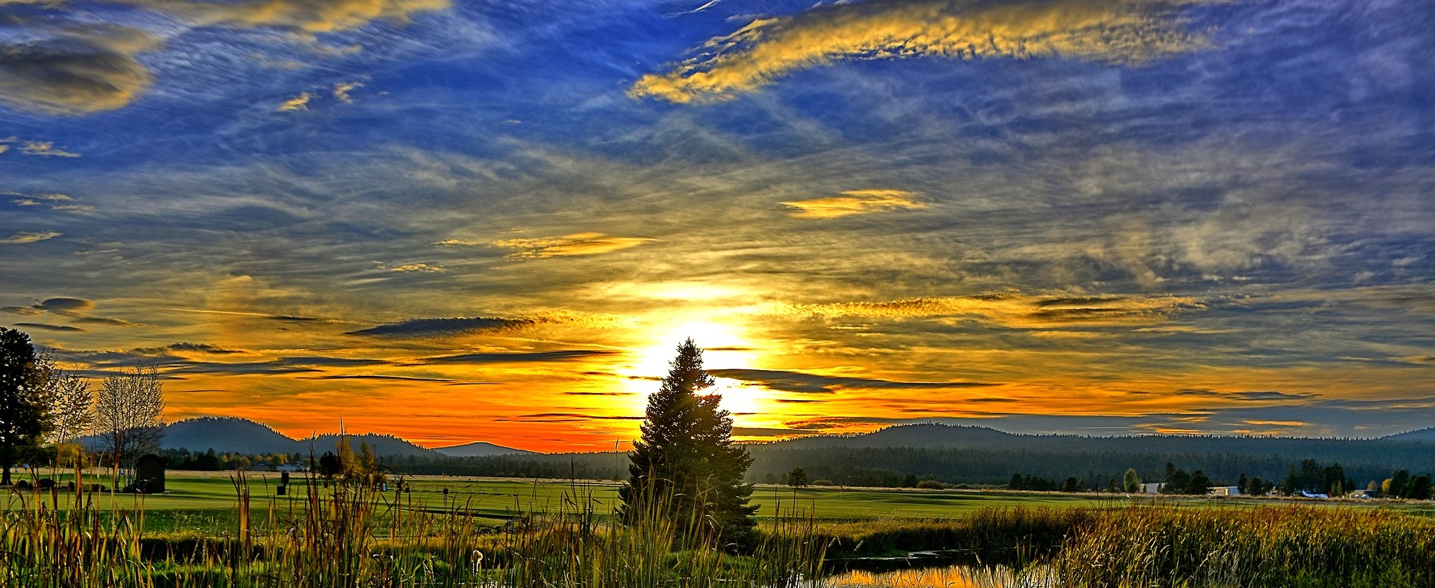 Landscape of sun setting behind rolling hills with green fields and a river in the foreground
