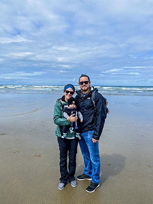 A mother and father, the mother with their baby strapped to her chest, posing at the beach.