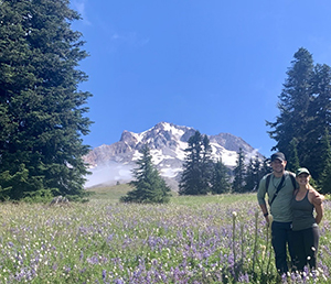 A man with his arm around a woman (both smiling) while on a hike on a sunny day with a mountain in the background.