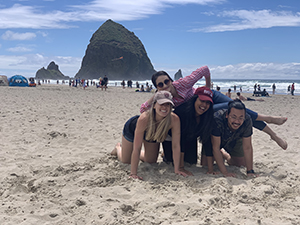 Two girls and one boy crouch down on the beach while a fourth woman lays on top of them, with Haystack Rock in the background.