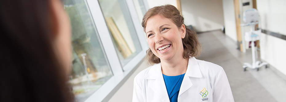 A woman wearing an OHSU branded white coat smiling while talking to someone in a hospital hallway.