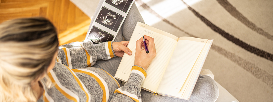 A pregnant woman writes in a notebook, with ultrasound images on her knee.