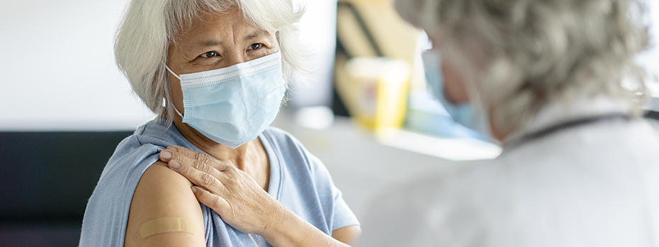 An older masked patient smiles and holds up her sleeve to show a Band-Aid she got after a vaccination.