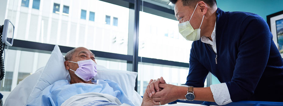 A doctor is comforting a patient who is recovering from surgery. The patient is lying in a hospital bed. They hold hands and smile at each other.