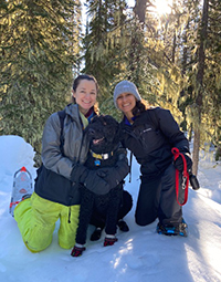 Two women kneeling down on either side of a dog in the snow on a sunny day.