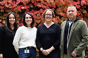 Four members of the I-CAN team smile in front of a wall of red ivy.