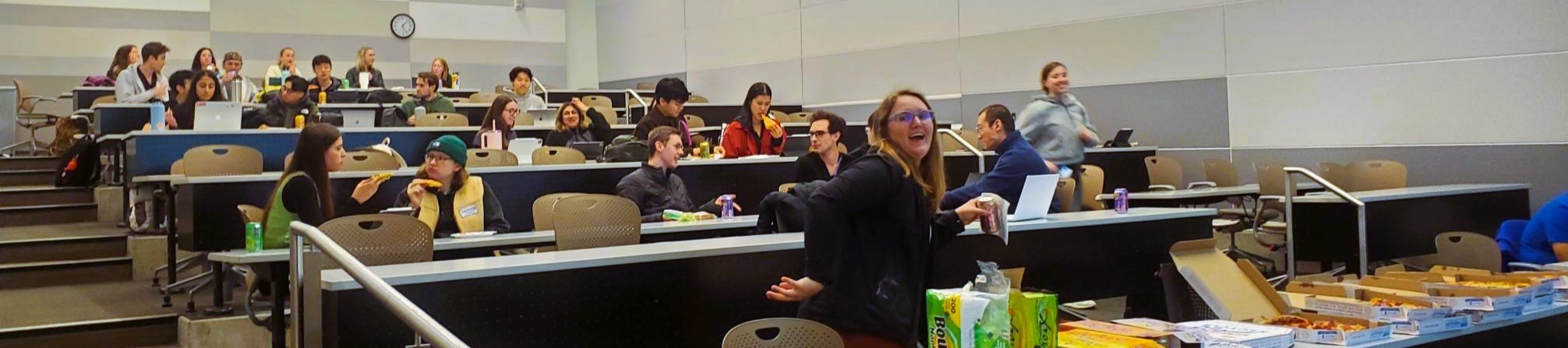 Students sitting in a tiered lecture hall. informally eating pizza and talking amongst themselves.