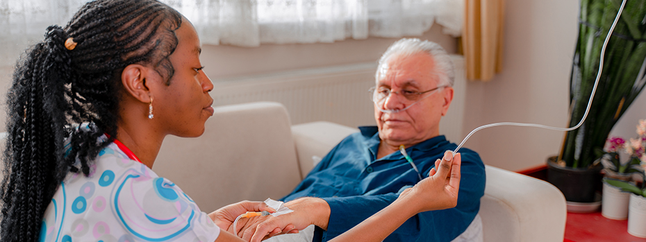 A patient lies on a couch while a nurse holds the patient’s hand with an IV needle already inserted and gets ready to attach an IV tube. 