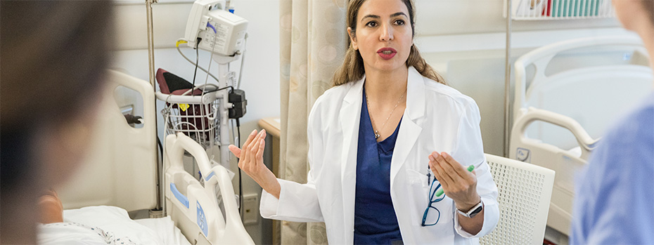 A health care professional in a white coat engages with colleagues in a hospital setting, demonstrating the kind of educational leadership role M.N.E. graduates can pursue. 