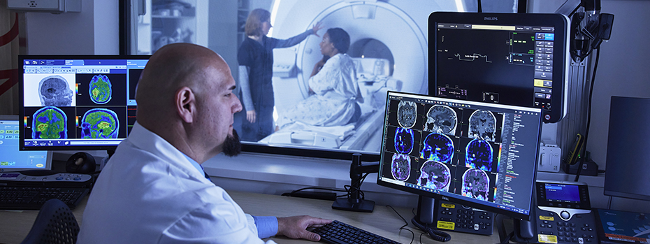 A doctor sits in front of two monitors showing digital scans and other data. A technologist and patient are visible through a window.