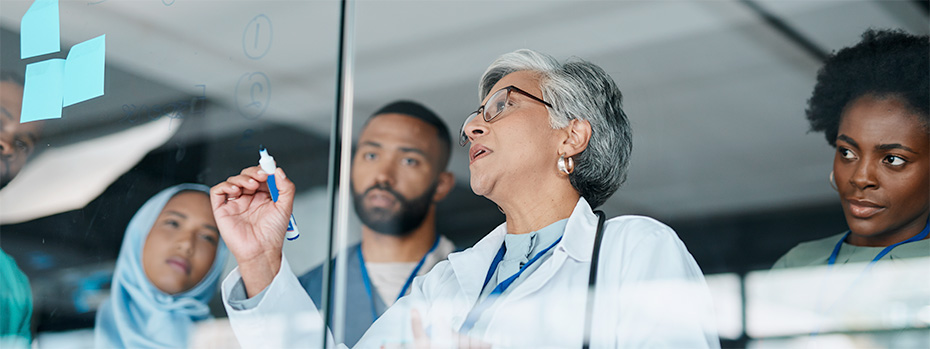 Research team members collaborate at a glass board where a nurse researcher writes notes as others observe the discussion.