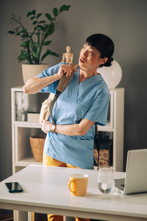 A healthcare asian woman in blue uniform is putting on a brown shoulder bag in their office space, which includes a desk, plants, and shelves, indicating readiness for the day.