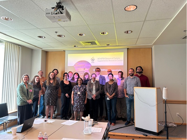 A group of 17 people stand closely together in a conference room in front of a screen with their presentation.