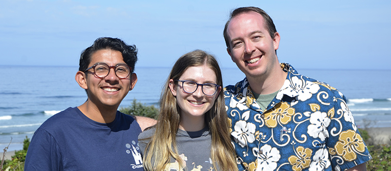 A woman standing between two men smiling with the ocean visible behind them on a sunny day.