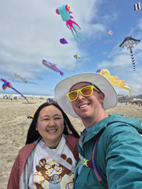 A woman and a man taking a selfie at the beach with kites flying in the background.