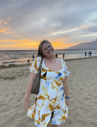 A woman standing at the beach smiling at sunset with the ocean behind her.