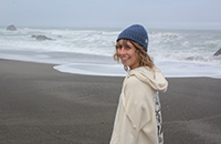 A woman smiling at the beach on a cloudy day with the ocean visible behind her.