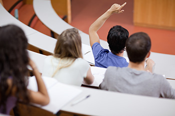 OHSU students sit in a classroom with hands raised, viewed from behind. One student in a blue shirt has their hand up to ask or answer a question.