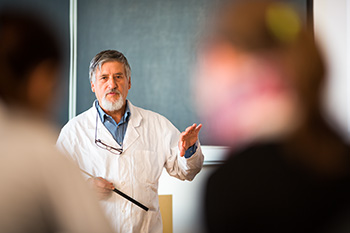 A medical educator in a white coat gestures while teaching, with students visible in the foreground out of focus.
