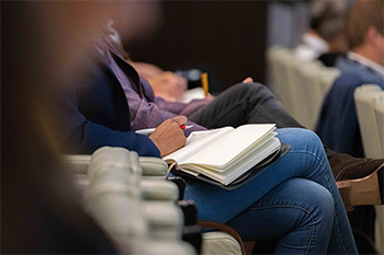 An OHSU educator takes notes in a notebook during a presentation, with colleagues in the background.