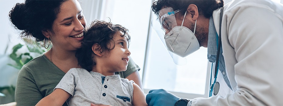A doctor wearing a mask and face shield places a bandage on child's arm while their mother holds them.