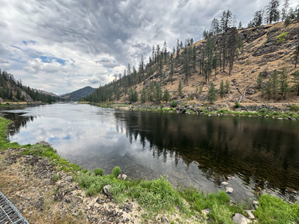Photograph of Clearwater River in Orofino Idaho. There is a calm river flowing alongside a rocky, tree-covered hillside under a cloudy sky. The scene features reflections of clouds on water surface, green vegetation near riverbank, and a mix of coniferous trees on the slope.